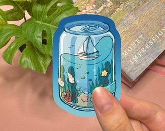 May include: A die-cut sticker shaped like a soda can featuring an underwater scene inside. The illustration shows a small white sailboat floating on the surface, with green seaweed, orange coral, seashells, and a yellow starfish at the bottom.
