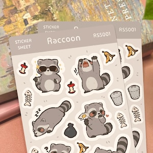 May include: A light gray sticker sheet labeled Raccoon RSS001 featuring cartoon raccoons in various poses, including wearing sunglasses, sleeping, and holding a flower. Additional small stickers include apple cores, fish bones, and trash cans.