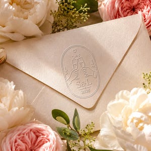 May include: A cream-coloured envelope featuring an embossed oval seal with a line art illustration of two hands clinking champagne glasses, the initials S&L, a small heart, and the year 2026, surrounded by white and pink peony flowers and greenery.