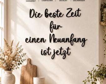 May include: Black wall-mounted lettering in a cursive script reading Die beste Zeit für einen Neuanfang ist jetzt. Above the text are four icons with labels: REGIONALE HANDARBEIT, PRÄZISE LASERTECHNIK, PERSÖNLICH GESTALTET, and BESONDERE GESCHENKIDEEN.