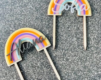 May include: Two rainbow cake toppers with wooden sticks. The rainbow is made of layered paper with orange, yellow, pink, purple, and blue colors. The name "Sofia" is written in black on the top topper and the name "Aliana" is written in black on the bottom topper.