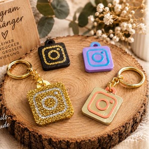 Könnte beinhalten: Vier quadratische Instagram-Logo-Schlüsselanhänger in Schwarz, Lila, Goldglitzer und Metallic-Gold mit pfirsichfarbenen Akzenten. Zwei Anhänger haben goldene Metallringe. Ein Holzschild zeigt den Text Instagram Anhänger, Kleiner Hingucker mit grosser Wirkung und Kleine Details, große Freude.