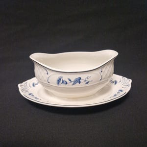 Könnte beinhalten: Weißes Soßenschiffchen und Untertasse mit blauem Blumendesign. Das Soßenschiffchen hat ein strukturiertes Muster und einen schwarzen Rand. Die Untertasse hat ein passendes blaues Blumendesign und einen schwarzen Rand.