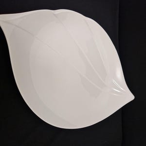 Könnte beinhalten: Eine weiße, blattförmige Servierschale mit glänzendem Finish. Die Schale hat ein glattes, geschwungenes Design mit subtilen linearen Details, ideal zum Servieren von Salaten oder Vorspeisen. Die Schale steht vor einem schwarzen Hintergrund.