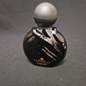 Könnte beinhalten: Eine schwarze, runde Parfümflasche mit kugelförmigem Verschluss. Die Flasche hat eine glänzende Oberfläche und der Text "EXPLOSIVE EAU DE TOILETTE NATURAL SPRAY" ist auf der Vorderseite aufgedruckt. Die Flasche steht auf dunklem Untergrund.