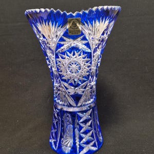 Könnte beinhalten: Eine blaue Kristallvase mit aufwendigen Stern- und geometrischen Mustern. Die Vase hat einen breiten, ausgestellten oberen Rand und ein detailliertes Design aus geschliffenem Glas. Ein kleines Etikett ist oben sichtbar. Die Vase dient wahrscheinlich dekorativen Zwecken.