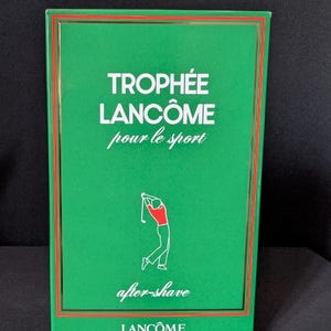 Könnte beinhalten: Grüne Schachtel mit Lancôme Trophée Aftershave. Die Schachtel hat einen roten Rand und trägt den weißen Schriftzug "Trophée Lancôme pour le sport". Eine Grafik eines Golfspielers in Rot und Weiß befindet sich darunter. Die Worte "after-shave" sind kursiv.