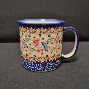 Könnte beinhalten: Eine Keramiktasse mit blauem Rand und Henkel. Die Tasse hat ein Blumenmuster mit roten, blauen und gelben Blumen auf cremefarbenem Hintergrund. Der Boden der Tasse hat einen blauen Zierrand.