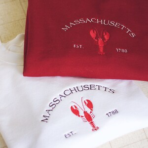 May include: Two folded sweatshirts, one red and one white, with embroidered logos. The logos feature a lobster and the text "MASSACHUSETTS EST. 1788".