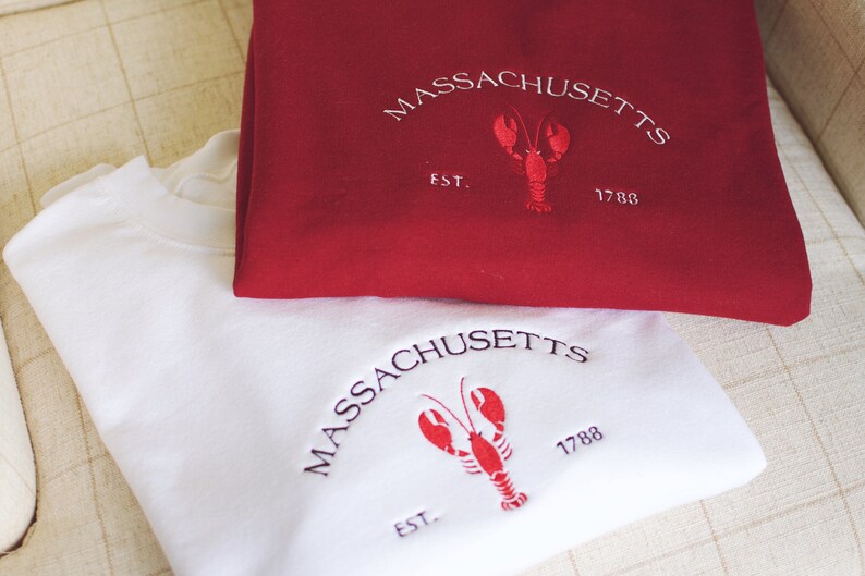 May include: Two folded sweatshirts, one red and one white, with embroidered logos. The logos feature a lobster and the text "MASSACHUSETTS EST. 1788".