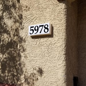May include: A rectangular white address plaque with the number 5978 in black, mounted on a textured beige wall. The wall has a red-brown trim and is partially shaded by tree branches.