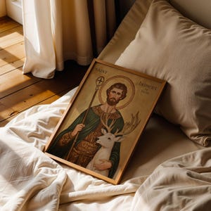 May include: A framed religious print featuring Saint Hubert of Li&egrave;ge. The artwork depicts a man with a beard and halo, holding a staff and embracing a white deer. The print is in a wooden frame and rests on a bed.
