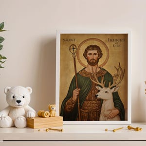 May include: A framed print featuring Saint Hubert of Li&egrave;ge, depicted with a deer. The artwork has a gold background and is framed in white. The text "SAINT HUBERT OF LIEGE" is visible. A white teddy bear and other decorative items are in the foreground.