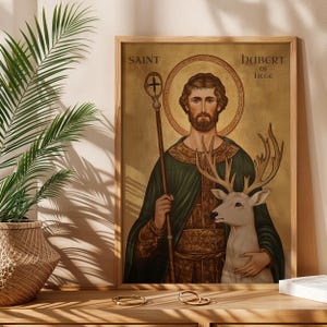 May include: Framed artwork featuring Saint Hubert of Li&egrave;ge, depicted with a halo, staff, and a white deer. The artwork has a gold background and is framed in a light wood. Text reads "Saint Hubert of Liege".