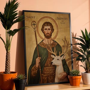 May include: Framed artwork depicting Saint Hubert of Li&egrave;ge. The image features a man with a halo, holding a staff and embracing a white deer. The artwork has a gold background and is framed in black. The text "SAINT" and "HUBERT OF LIEGE" are visible.