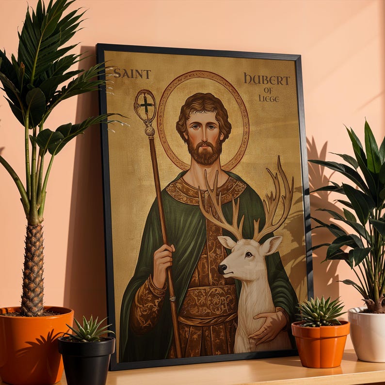 May include: Framed artwork depicting Saint Hubert of Li&egrave;ge. The image features a man with a halo, holding a staff and embracing a white deer. The artwork has a gold background and is framed in black. The text "SAINT" and "HUBERT OF LIEGE" are visible.