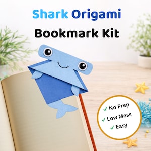 May include: A blue origami shark bookmark kit. The bookmark is made of blue paper and is inserted into an open book. The text "Shark Origami Bookmark Kit" is at the top of the image. The kit is described as "No Prep", "Low Mess", and "Easy".