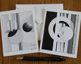 May include: Two black and white art cards with geometric designs, accompanied by a white envelope. The cards feature circles, lines, and shading. A black pen rests below the cards on a wooden surface.