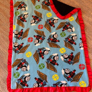 May include: A baby blanket featuring a repeating pattern of a cartoon train on a light blue background. The blanket has a red ruffled satin border and a black textured backing.