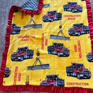 May include: A yellow fleece baby blanket with a red satin trim. The blanket features a repeating pattern of red dump trucks with the words "Construction Ahead" printed on the blanket.