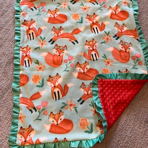 May include: A soft, blue and red baby blanket with a fox print. The blanket has a green satin trim and a soft, textured back.