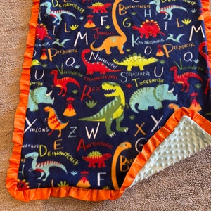 May include: A blue fleece blanket with an orange ruffle border. The blanket features a dinosaur alphabet pattern with colorful dinosaurs and letters. The back of the blanket is a light blue minky fabric.