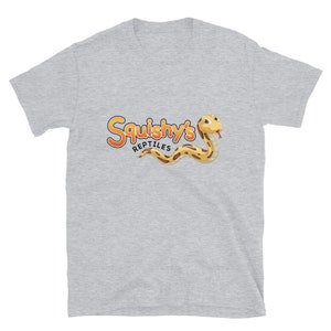 Op de afbeelding: Lichtgrijs t-shirt met de tekst "Squishy's Reptiles" in oranje en geel, en een cartoon slangenillustratie. De slang is geel en bruin met een rode tong.