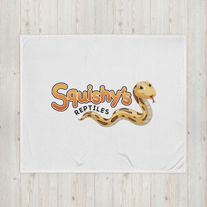 Könnte beinhalten: Eine weiße Decke mit einer Cartoon-Schlange und dem Text "Squishy's Reptiles" in Orange und Gelb.