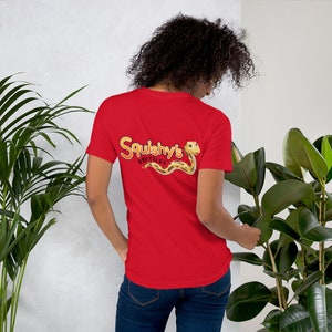Op de afbeelding: Een rood t-shirt met de tekst "Squishy's Reptiles" in een gele en bruine letter, met een cartoon slang. Het shirt heeft korte mouwen en is gemaakt van een zacht materiaal. Het ontwerp staat op de achterkant van het shirt.