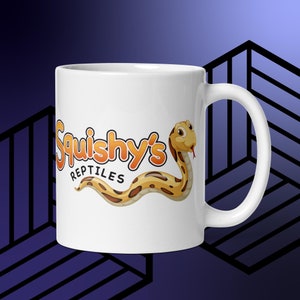 Könnte beinhalten: Weiße Keramik-Tasse mit dem Text "Squishy's Reptiles" in Orange und einer Cartoon-Schlangen-Illustration. Die Schlange ist gelb und braun mit roter Zunge. Die Tasse hat einen gebogenen Henkel.