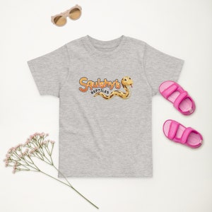Peut inclure: Un t-shirt gris à manches courtes avec un serpent de dessin animé et le texte "Squishy's Reptiles" en orange et jaune. Le t-shirt est sur un fond blanc avec des sandales roses et une brindille de fleurs blanches.