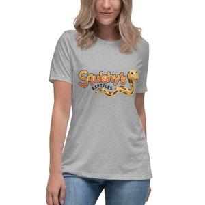 Può includere: T-shirt grigio chiaro con la scritta "Squishy's Reptiles" in arancione e giallo, e un'illustrazione di un serpente dei cartoni animati. Il serpente è giallo e marrone. La maglietta è realizzata in un materiale morbido.