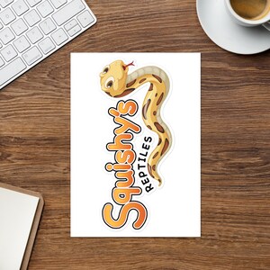 Könnte beinhalten: Ein Aufkleber mit einer Cartoon-Schlange und dem Text "Squishy's Reptiles" in Orange und Gelb.