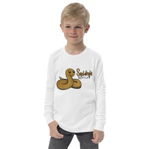 Könnte beinhalten: Weißes Langarmshirt mit einer Cartoon-Schlangenillustration und dem Text "Squishy's Reptiles" in Braun. Die Schlange ist hellbraun mit einem lächelnden Gesicht. Das Shirt wird von einer jungen Person getragen.
