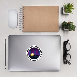 May include: A round sticker with a black border and white background. The sticker features a purple and blue aurora borealis with a yellow sun setting behind a dark silhouette of trees. The text "SPACE WEATHER WATCH" is written in a black circle around the image.