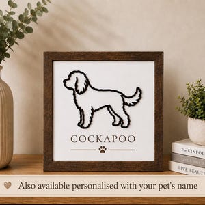 May include: A square wall sign with a dark brown wooden frame and a white background. The sign features a black outline of a Cockapoo dog, the text "COCKAPOO" in black, and a small black paw print icon centred below the dog silhouette.