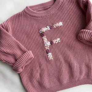 May include: A dusky pink knitted sweater with a ribbed texture. The front of the sweater is adorned with a floral embroidered "F", using white, pink, and purple flowers. The sleeves are cuffed, and the neckline is a classic crew neck.