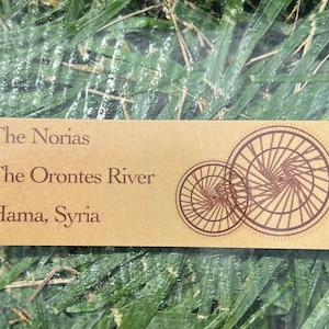 Könnte beinhalten: Eine rechteckige, hellbraune Karte mit dem Text "The Norias", "The Orontes River" und "Hama, Syrien" in braunen Buchstaben. Auf der rechten Seite befinden sich zwei sich überlappende Kreisdesigns. Die Karte liegt auf grünem Gras.