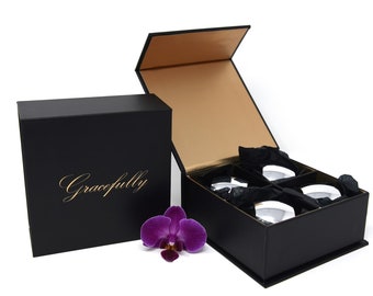 Op de afbeelding: Een zwarte geschenkdoos met het woord "Gracefully" gedrukt in gouden letters. De doos is open en onthult vier kleine, ronde, witte containers erin. De containers zijn genesteld in zwart tissuepapier. Een paarse orchidee bevindt zich op de voorgrond.