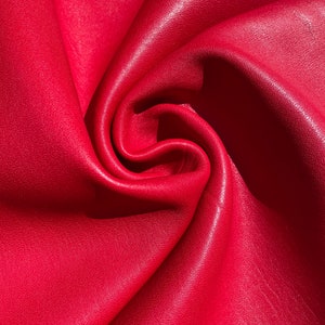May include: Close-up of a smooth, red leather fabric with a textured surface. The fabric is folded and draped, creating a soft, flowing look.