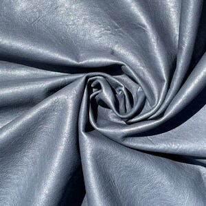 May include: Close-up of a smooth, grey leather fabric with a subtle sheen. The fabric is folded and draped, creating soft folds and curves.