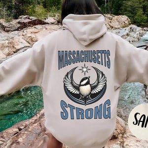 May include: Sand-colored hoodie with the words "MASSACHUSETTS STRONG" in blue lettering. A graphic of a bird with outstretched wings is centered between the words. The hoodie has a hood and long sleeves. A circular "SAND" label is in the lower right corner.