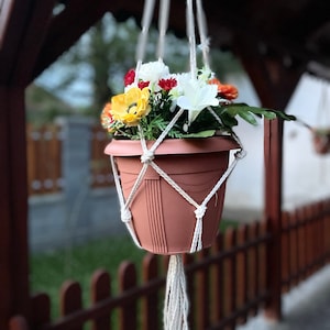 Puede incluir: Una maceta colgante de color terracota llena de coloridas flores artificiales. La maceta está suspendida por un colgador de plantas de macramé blanco. Las flores incluyen flores amarillas, rojas, blancas y naranjas, con follaje verde.