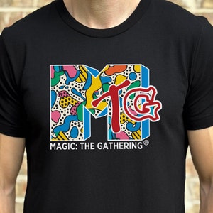 May include: Black t-shirt with a colorful graphic of the Magic: The Gathering logo. The logo is a stylized "M" with a "TG" inside.