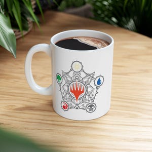 May include: White ceramic mug with a black handle. The mug features a graphic of a five-pointed star with five symbols inside. The symbols are a white sun, a green leaf, a blue water drop, a red flame, and a black skull.