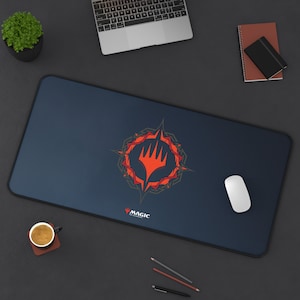 May include: A dark blue mousepad with a red and gold Magic: The Gathering logo. The logo features a five-pointed star with a stylized hand inside. The mousepad is on a gray surface with a cup of coffee, pencils, and a computer mouse.