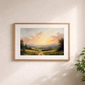 May include: A framed landscape painting depicts a serene countryside scene. The artwork features a dirt path winding through green fields towards distant hills under a sunset sky with orange and yellow hues. The frame is made of light brown wood.