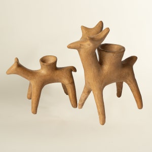 Puede incluir: Tres esculturas de ciervos de cerámica, cada una con una parte superior hueca, están dispuestas sobre un fondo blanco. Los ciervos están todos mirando hacia el lado derecho de la imagen. El ciervo más grande está en la parte posterior, con un ciervo más pequeño delante de él, y el ciervo más pequeño está en la parte delantera. Los ciervos son todos de color marrón claro.