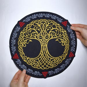 May include: A circular black patch with a gold Celtic tree of life design. It is encircled by a silver leaf border and red knotwork accents. The patch is held by two hands.