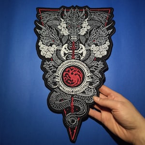 May include: A black and white embroidered patch featuring a three-headed dragon with red eyes and a red dragon symbol in the center. The dragon is surrounded by a circle with a white background and a black border. The patch is shaped like a triangle with a pointed bottom.